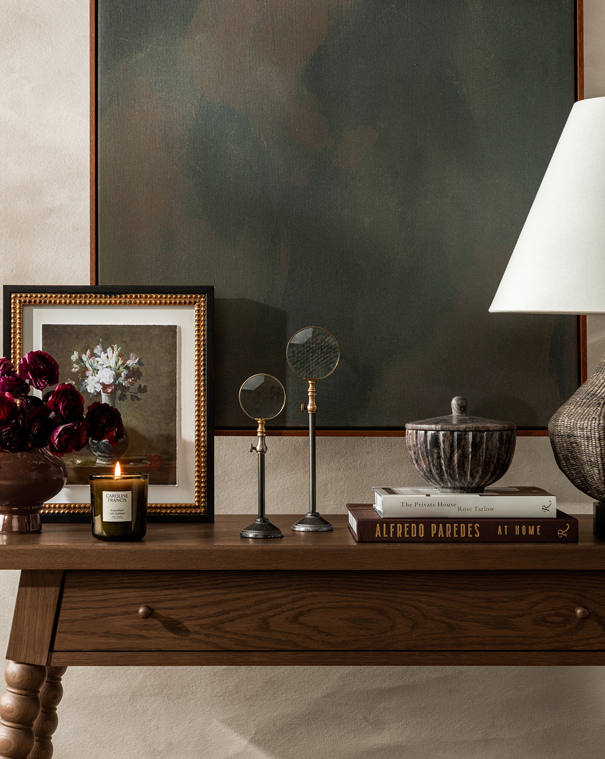A wooden table holds a framed floral painting, an Antique Flower Vase with dark red flowers, a lit candle, two magnifying glasses, a lidded bowl, stacked books, and a sculpted lamp, with vintage wall art displayed above by McGee & Co.