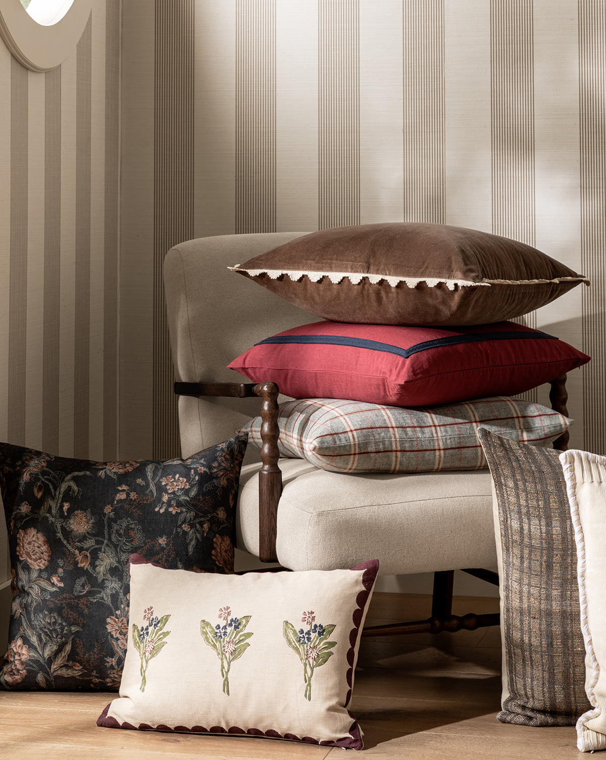 A beige armchair with assorted pillows, including the vintage-inspired Hestercombe Pillow Cover, sits next to a striped throw blanket and a decorative pillow in a cozy, softly lit room, by McGee & Co.