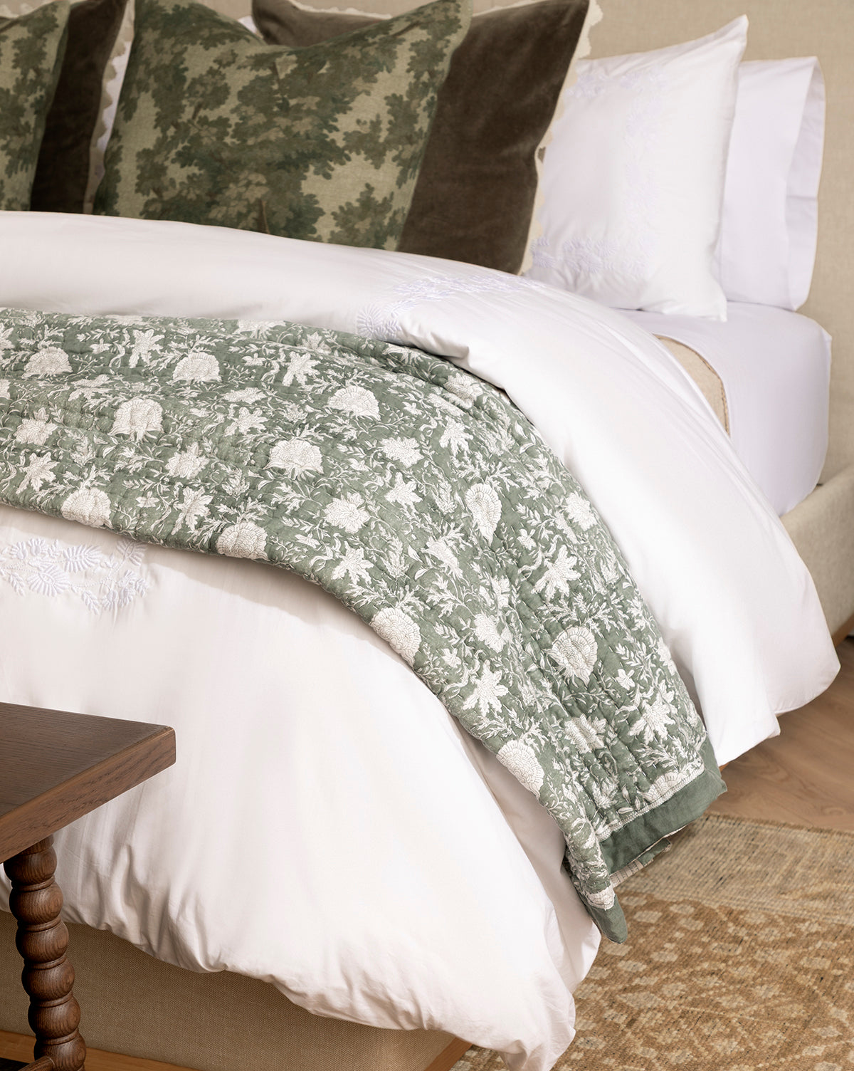A neatly made bed with white sheets, two green and brown decorative pillows, and the Audrey Sage Block Print Quilt with ruffle edging folded at the foot, by McGee & Co. A wooden side table and woven rug are partially visible.