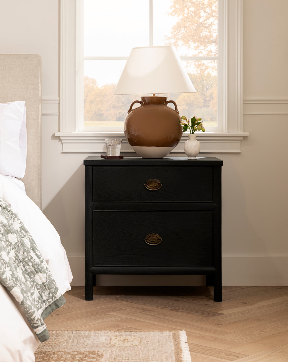 A black nightstand with two drawers sits by the bed. On top are an Amphora Table Lamp, a glass of water, a small plant, and a book. Natural light streams through a window in the background, by McGee & Co.