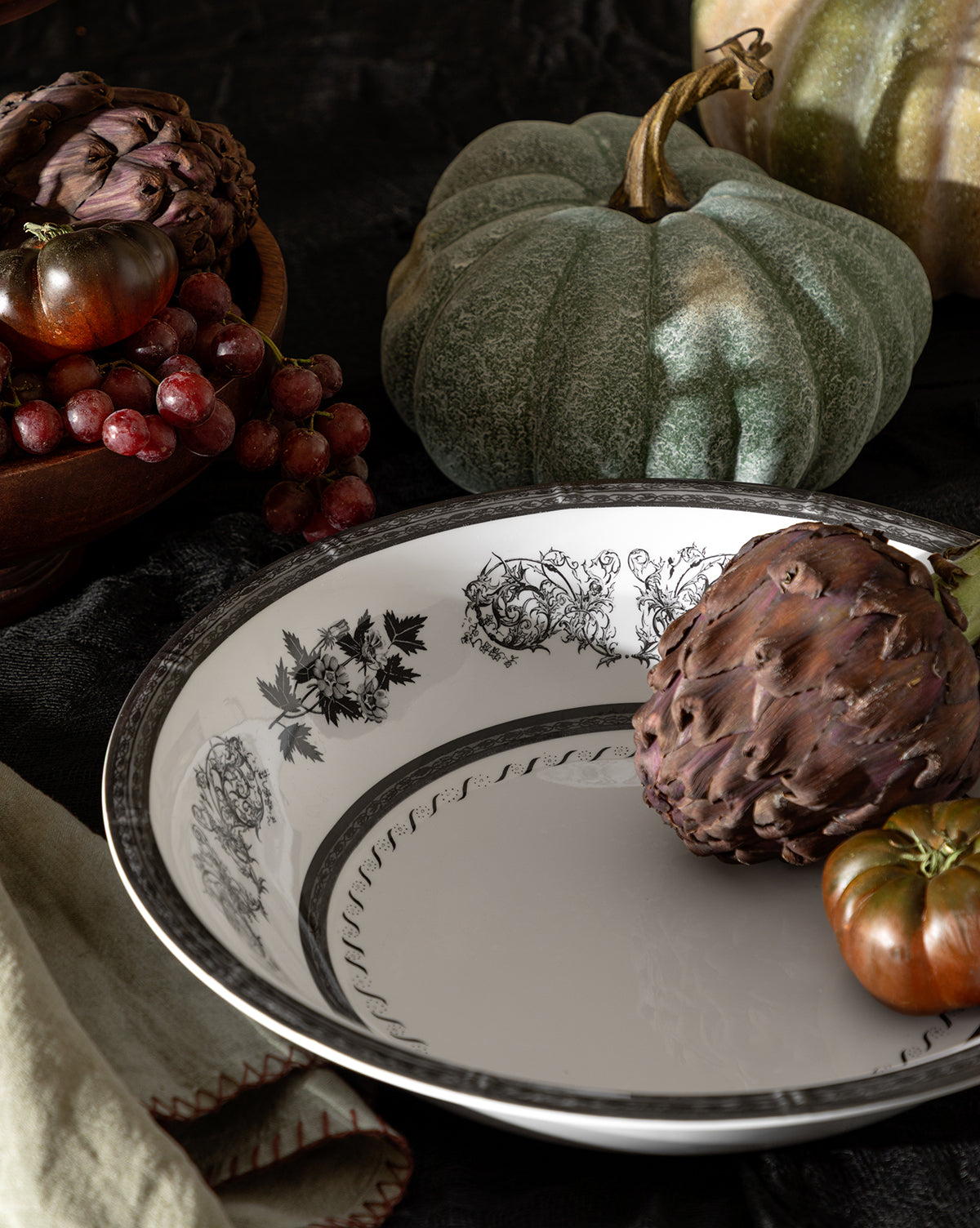The Calanthe Melamine Serving Bowl, adorned with black floral designs, sits on a table among an artichoke, green pumpkin, grapes, and tomatoes for a rustic autumnal display, by McGee & Co.