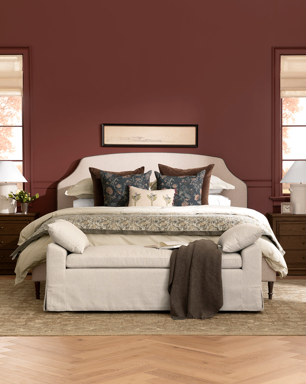 A cozy bedroom with a king-sized bed, layered pillows, and the sustainable Locester Throw. A beige bench sits at the foot of the bed, wooden nightstands with lamps flank each side, and a brown accent wall features framed artwork above by McGee & Co.
