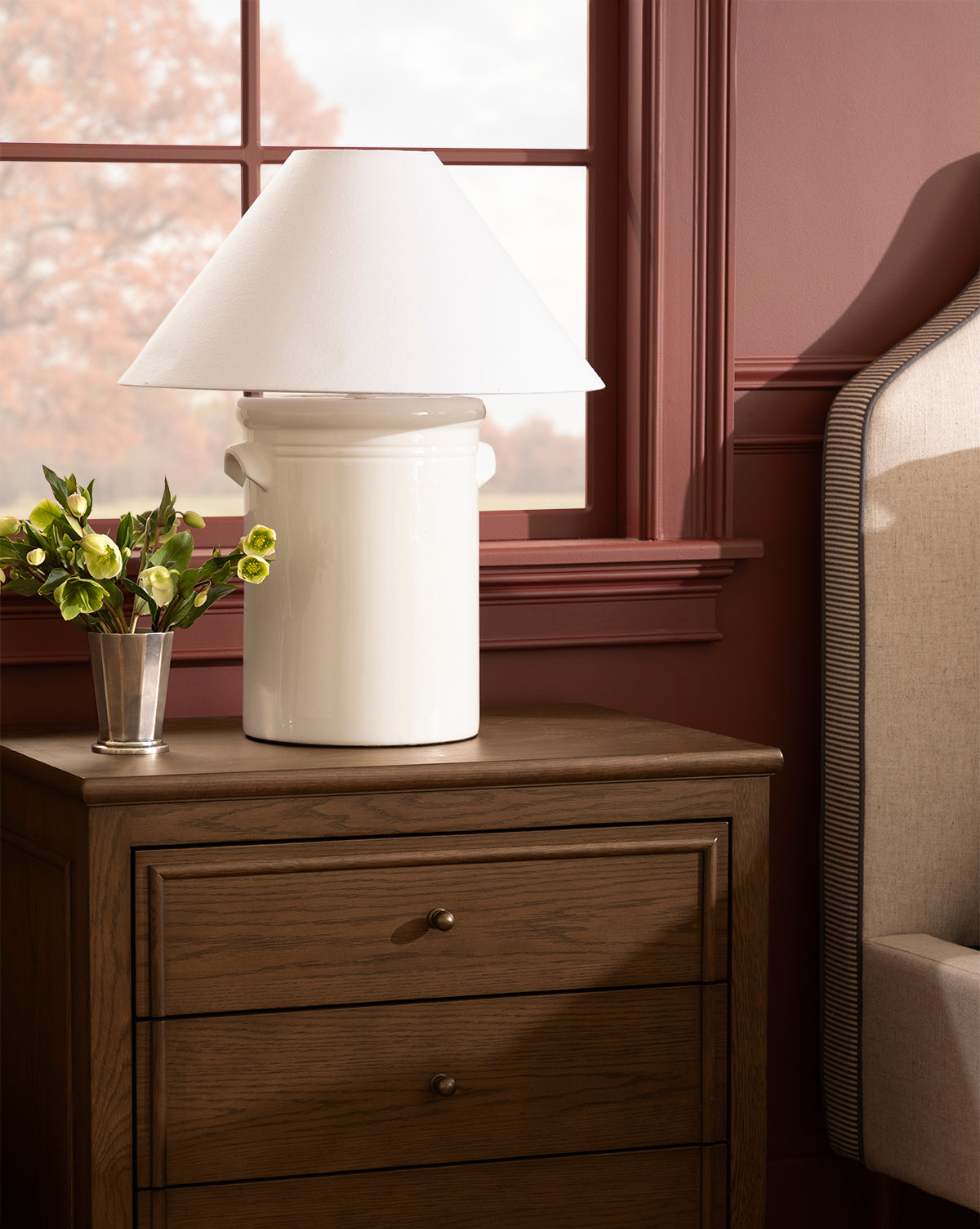 The Arles Table Lamp elevates your wooden nightstand with its elegant ceramic base and conical shade, complementing a small vase of flowers, an upholstered bed, and a window with reddish-brown trim, by McGee & Co.