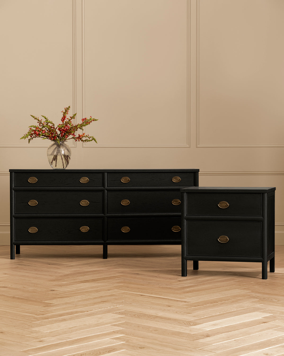 Two black dressers, styled like the Edward Nightstand with gold oval handles, stand on a light herringbone wood floor against a beige paneled wall; the larger dresser displays a clear vase with red and green flowers by McGee & Co.