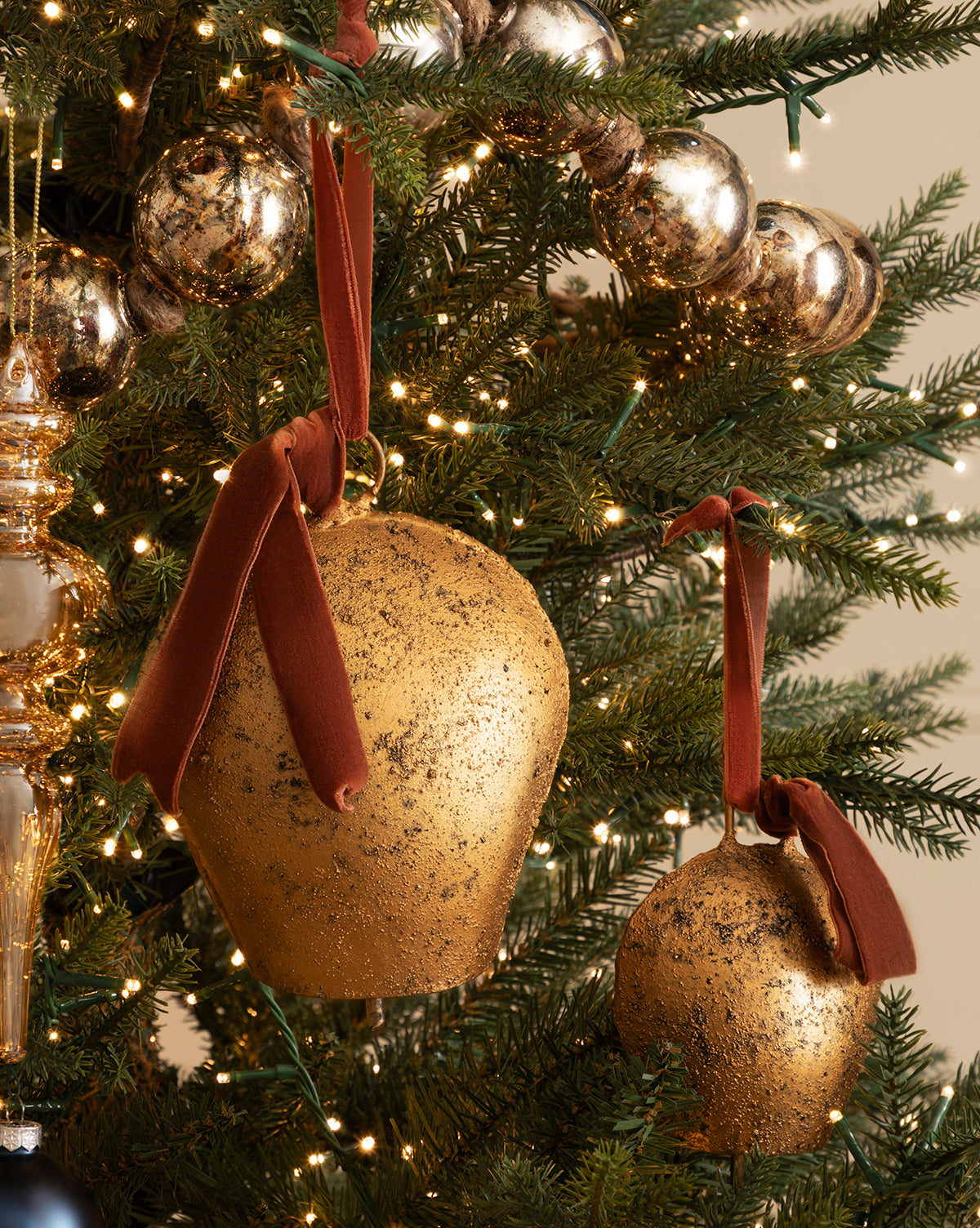 Two Antique Gold Metal Bell with Velvet Ribbon ornaments hang on a Christmas tree decorated with sparkling lights and metallic garland, bringing timeless holiday charm, by McGee & Co.