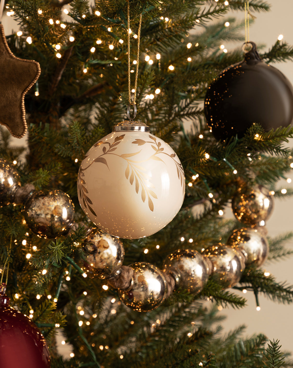 A close-up of a decorated Christmas tree with the 4" Hand-Painted Vine Glass Bauble, beaded ornaments, metallic garland, Fish with Sequins Ornaments, a black ornament, and glowing string lights for festive holiday décor, by McGee & Co.