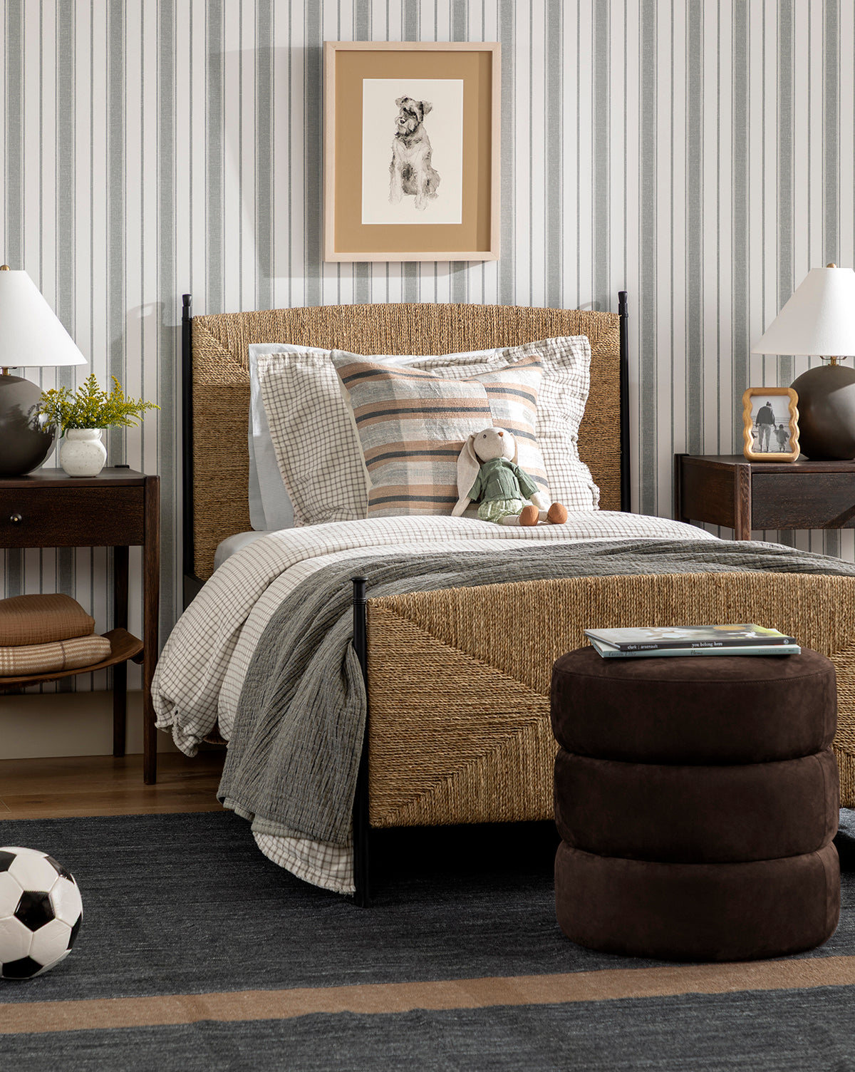 A cozy bedroom with a woven bed, striped wall, framed dog sketch, two bedside tables with lamps, plush rabbit toy on the bed, Channel Ottomans at the foot, and a soccer ball on a dark quilted rug by McGee & Co.