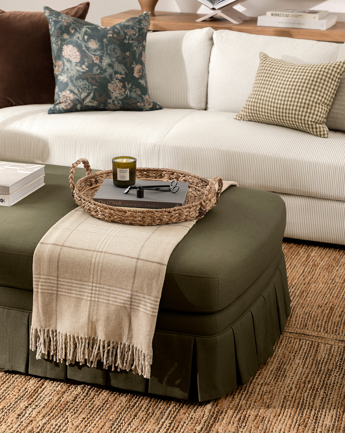 A cozy living room has a white sofa with patterned pillows, a green ottoman with a beige plaid throw, and the Salter Woven Tray displaying a candle, book, and glasses. A textured rug completes the inviting space, by McGee & Co.