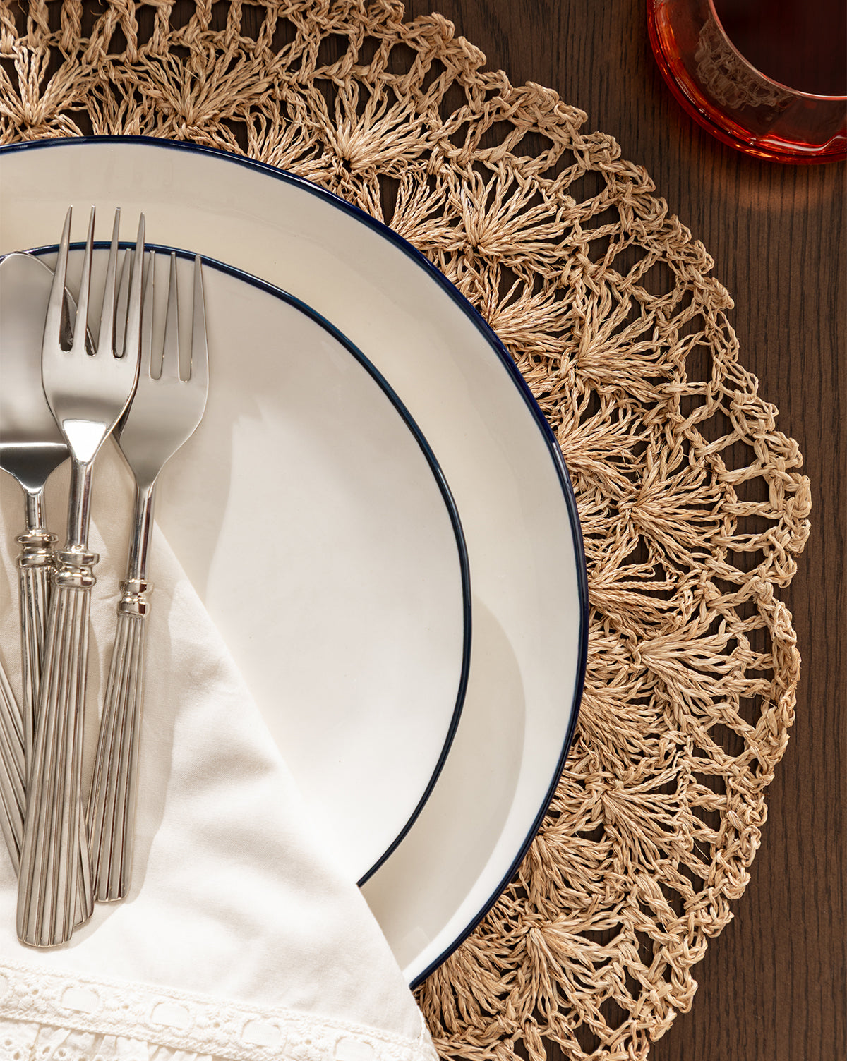 Overhead view of two stacked white plates with dark rims on the Hermione Placemat, topped with a folded napkin and three silver forks, next to a glass containing a reddish drink by McGee & Co.