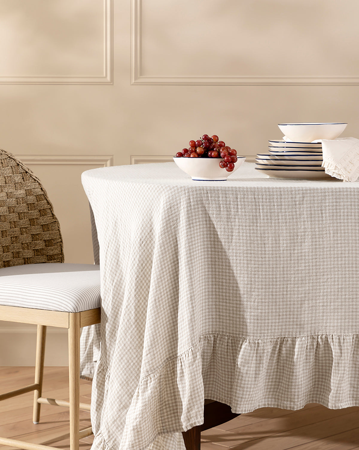 A round table draped with a beige Catania Linen Ruffle Tablecloth in gingham is set with stacked white plates, a napkin, and a bowl of red grapes. A woven chair with a cushioned seat sits beside this inviting setup, by McGee & Co.