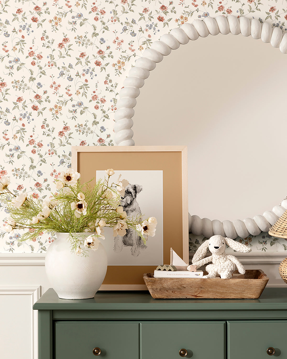 A charming tabletop features the Isla Terracotta Vase with flowers, a framed dog sketch, a toy sailboat and stuffed bunny on a wooden tray, all set before a round beaded mirror and floral wallpaper for delightful home decor, by McGee & Co.