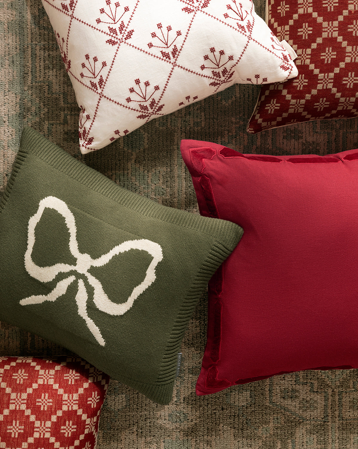 Four pillow covers are arranged on a rug, featuring festive holiday layering in red, white, and green. A green pillow with a white bow stands out among Bella Pillow Covers decorated with geometric and floral patterns, by McGee & Co.