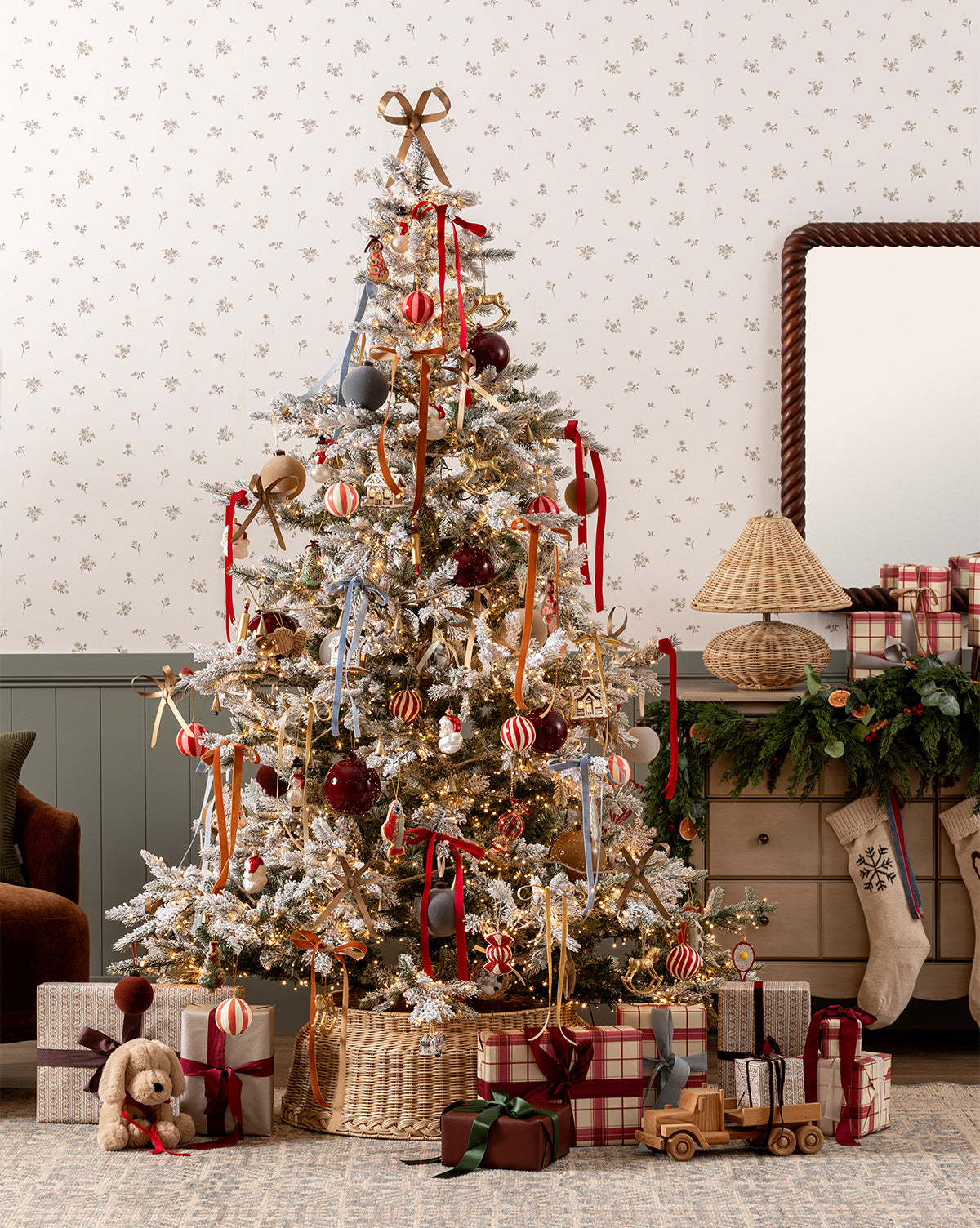 The Magical Wonderland Tree stands in a cozy room with red, gold, and striped ornaments. Wrapped presents, a toy teddy bear, and a wooden truck sit beneath. Garland and a lamp on the dresser complete the festive holiday decor, by McGee & Co.