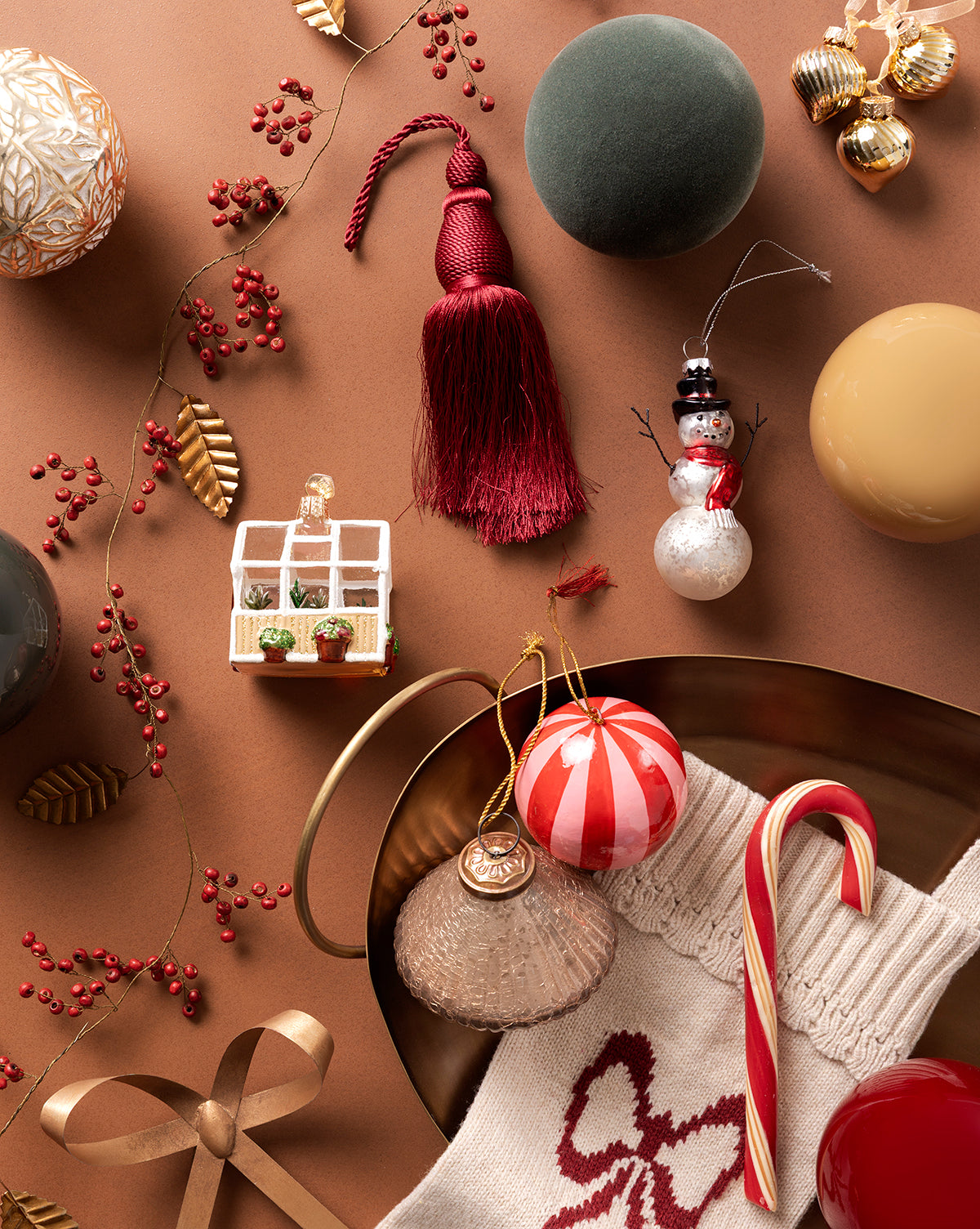 A festive flatlay with holiday ornaments, a red tassel, candy cane, knit stocking, gold ribbon, faux berries, and a red-and-white bauble from the 4" Assorted Flocked Baubles (Set of 4), all on a brown background, by McGee & Co.