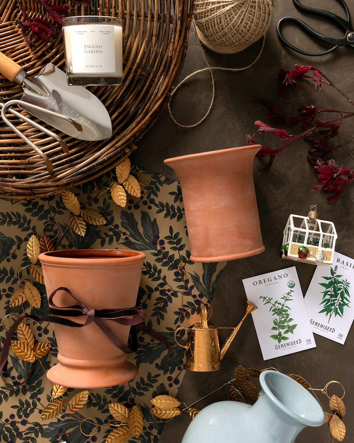 A flat lay of gardening items, including terracotta pots, a gold Watering Can Ornament with a copper finish, a white trowel, scissors, seed packets, twine, a candle, and leafy decorative paper on a brown surface, by McGee & Co.