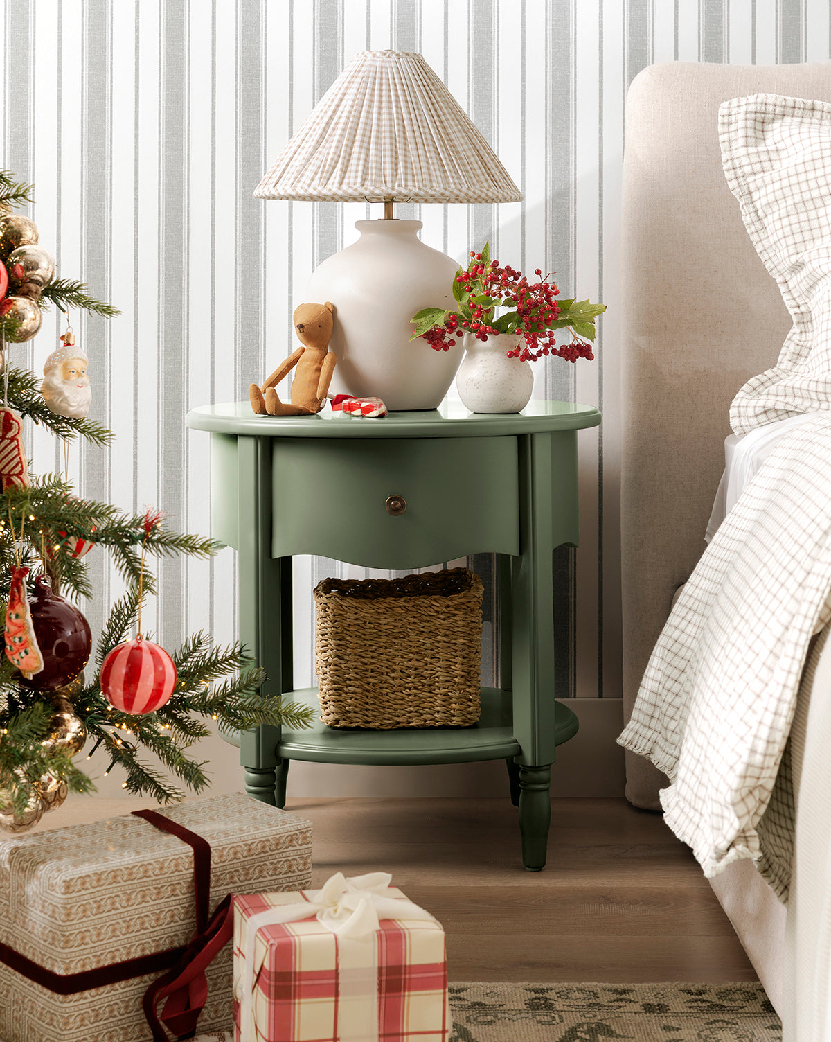 A green round nightstand with a lamp, teddy bear, and festive decor—including the Lyra Vase—sits beside a beige bed. A basket is on the lower shelf. A Christmas tree and wrapped gifts stand in front of striped wallpaper, by McGee & Co.