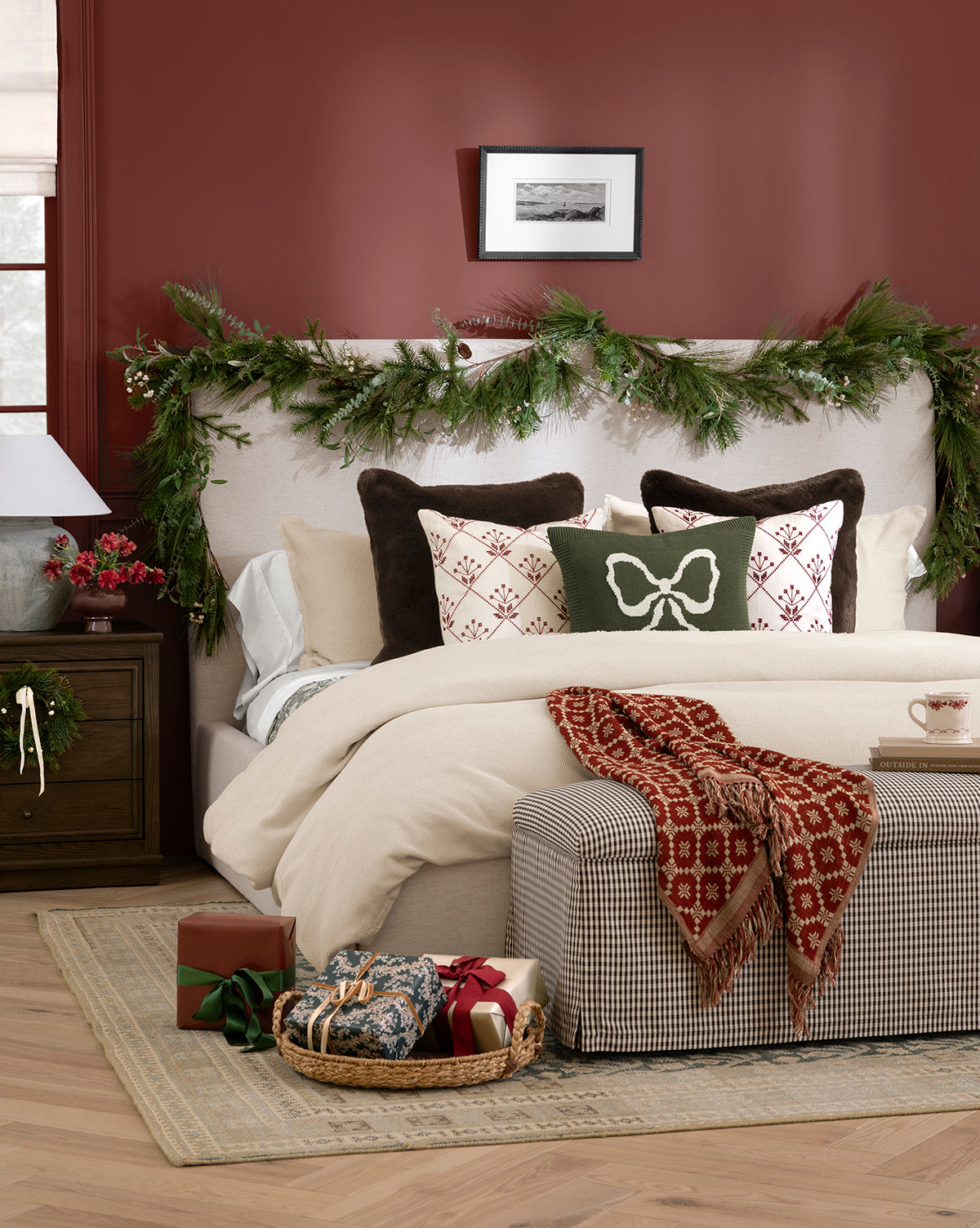 A cozy bedroom dressed for the holidays features pine garlands on the headboard, festive pillows, wrapped gifts, a basket at the foot of the bed, and a Jude Reversible Woven Throw draped across crisp bedding against a rich red accent wall, by McGee & Co.