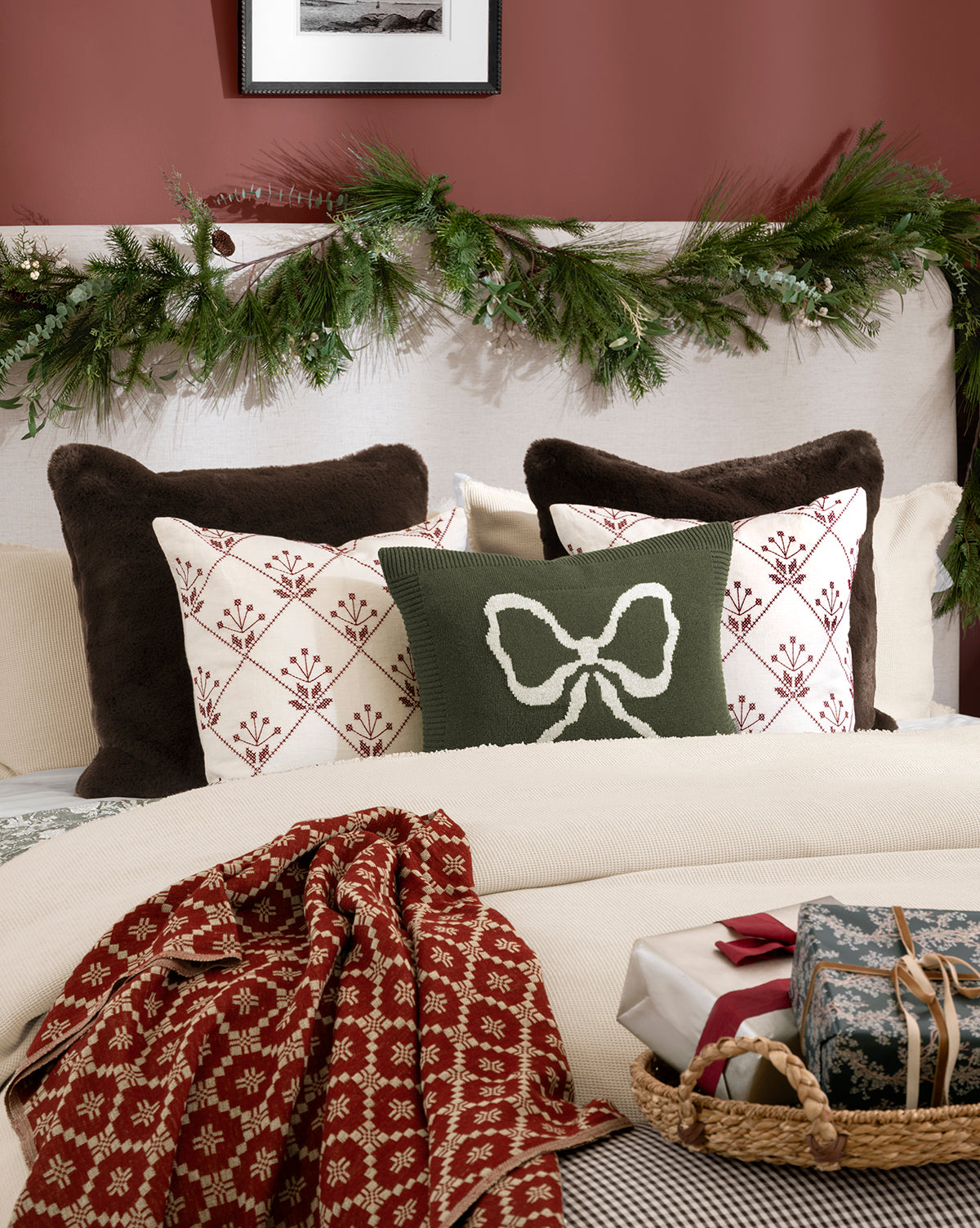 A cozy bed features brown and white pillows, a green pillow with a white bow, the McCallister Pillow Cover, a red patterned throw, gift boxes, and festive greenery garland over a white headboard—ideal for holiday home decor, by McGee & Co.
