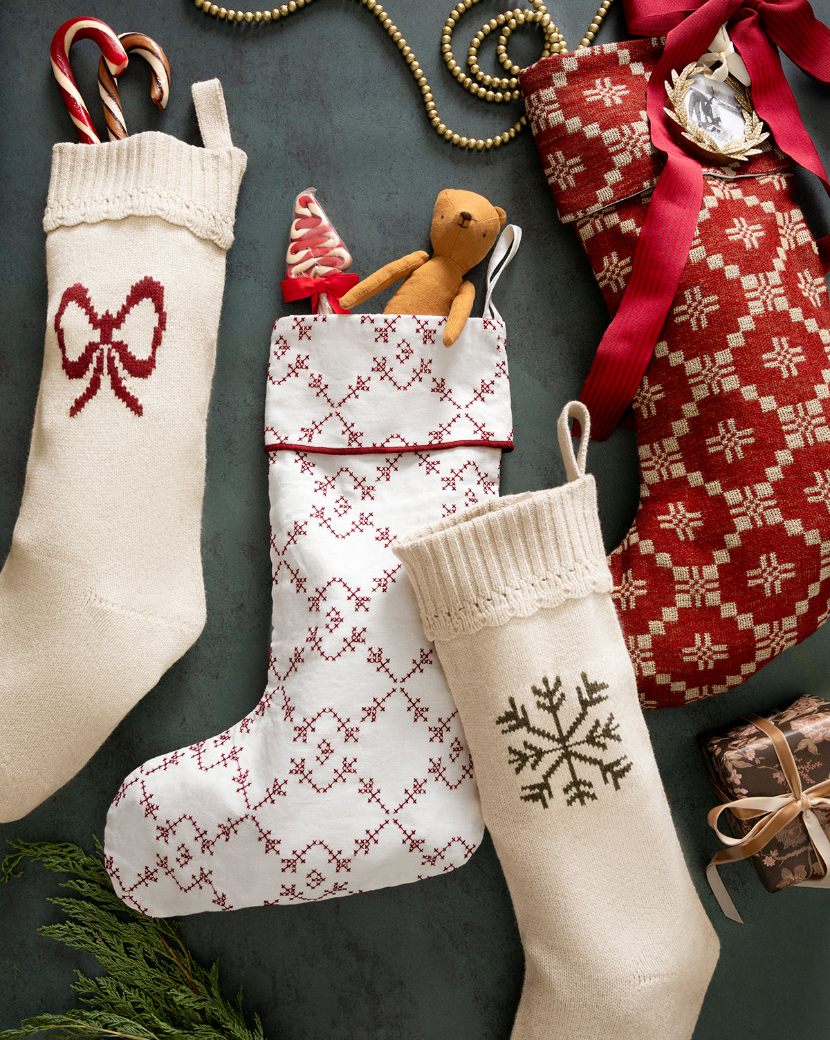 The Bella Scalloped Stocking with Bow is filled with candy canes and a teddy bear, arranged alongside festive beads and a wrapped gift, creating charming holiday mantel decor on a dark surface, by McGee & Co.