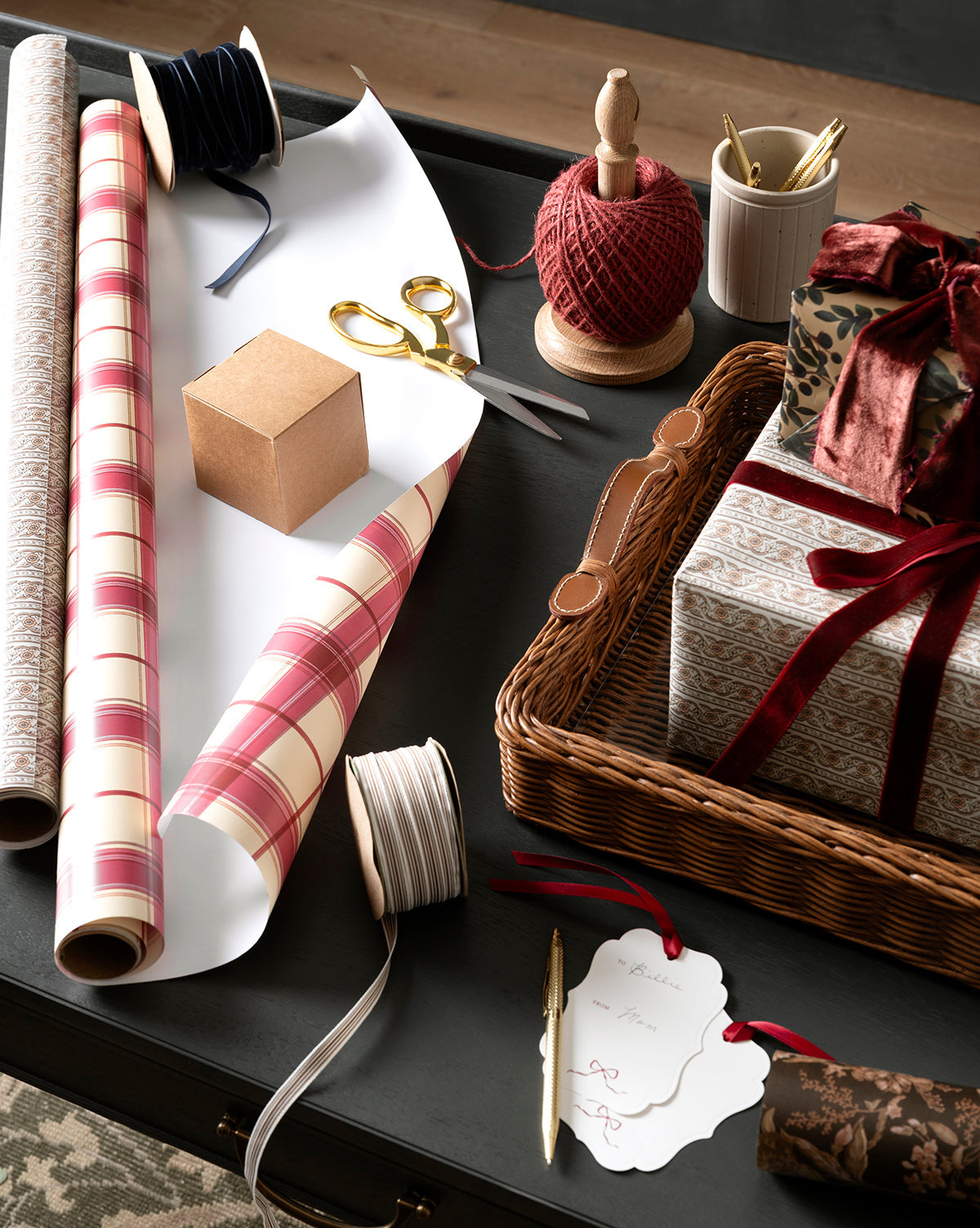 A gift-wrapping setup featuring patterned paper rolls, scissors, a 3/8" Oatmeal & Brown Woven Grosgrain Stripe Ribbon, twine, blank gift tag, pen, wicker basket with wrapped gifts, and an unwrapped brown box—perfect for holiday decorations, by McGee & Co.