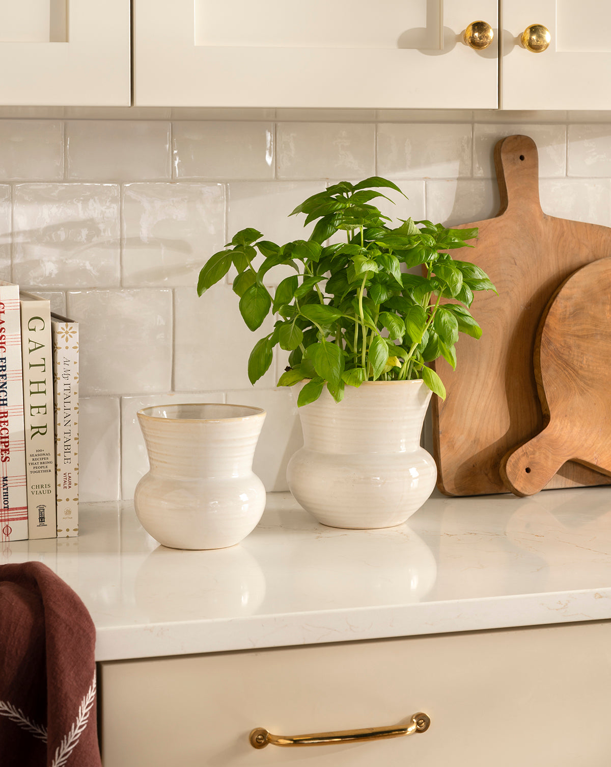 A kitchen countertop features two white ceramic Colchester Planters—one with a lush basil plant—alongside wooden cutting boards, cookbooks, and gold-accented white cabinets for stylish home decor, by McGee & Co.