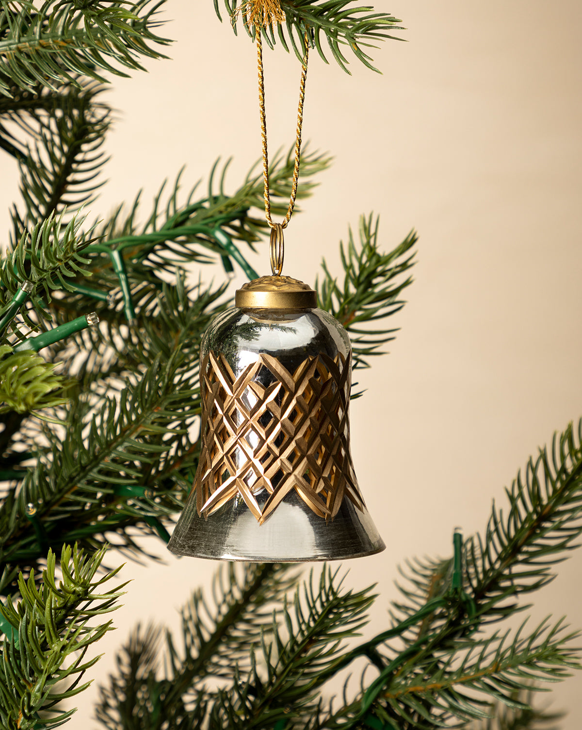 The Hand-Painted Mercury Glass Bell Ornament, featuring a gold lattice design, hangs from a green Christmas tree branch against a beige background, bringing elegant holiday décor to your festive setting, by McGee & Co.