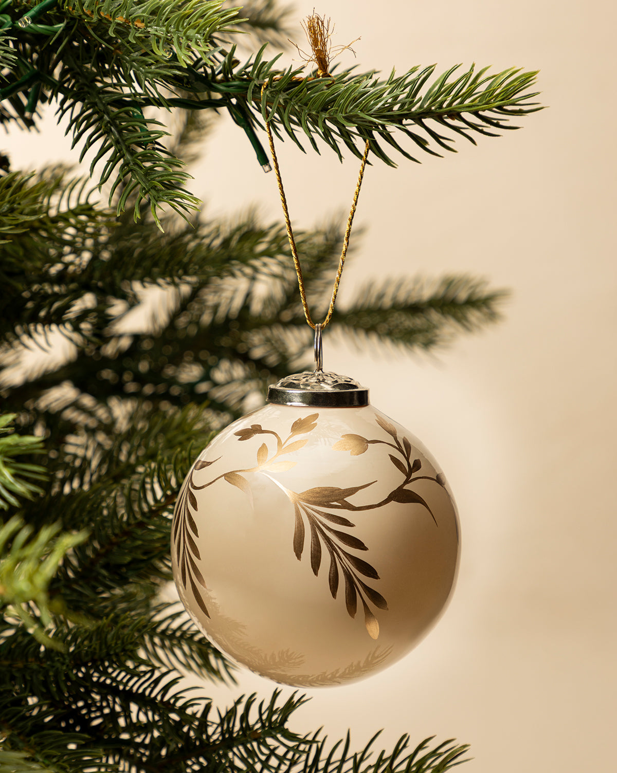 The 4" Hand-Painted Vine Glass Bauble, featuring gold and white beads in a leafy design, hangs from a green pine branch and brings elegant style to your holiday decor against a neutral backdrop, by McGee & Co.