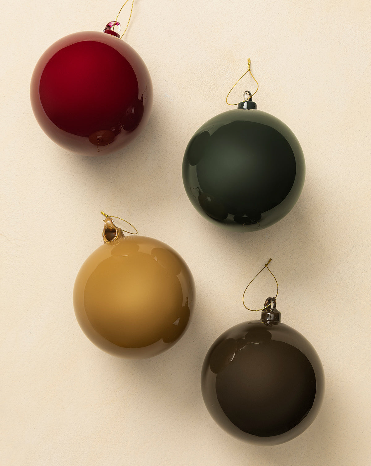 The 5" High Gloss Glass Baubles (Set of 4) in red, dark green, gold, and brown are arranged on a light beige surface. Each features a gold hanging string, making them perfect for holiday decorating, by McGee & Co.