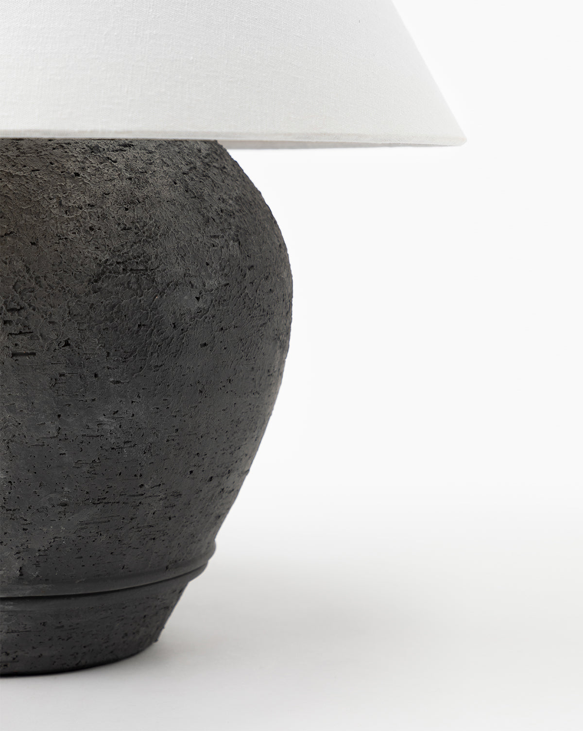 A close-up of the Abbott Ceramic Table Lamp featuring a textured black ceramic round base and a white linen shade, set against a plain white background, by McGee & Co.