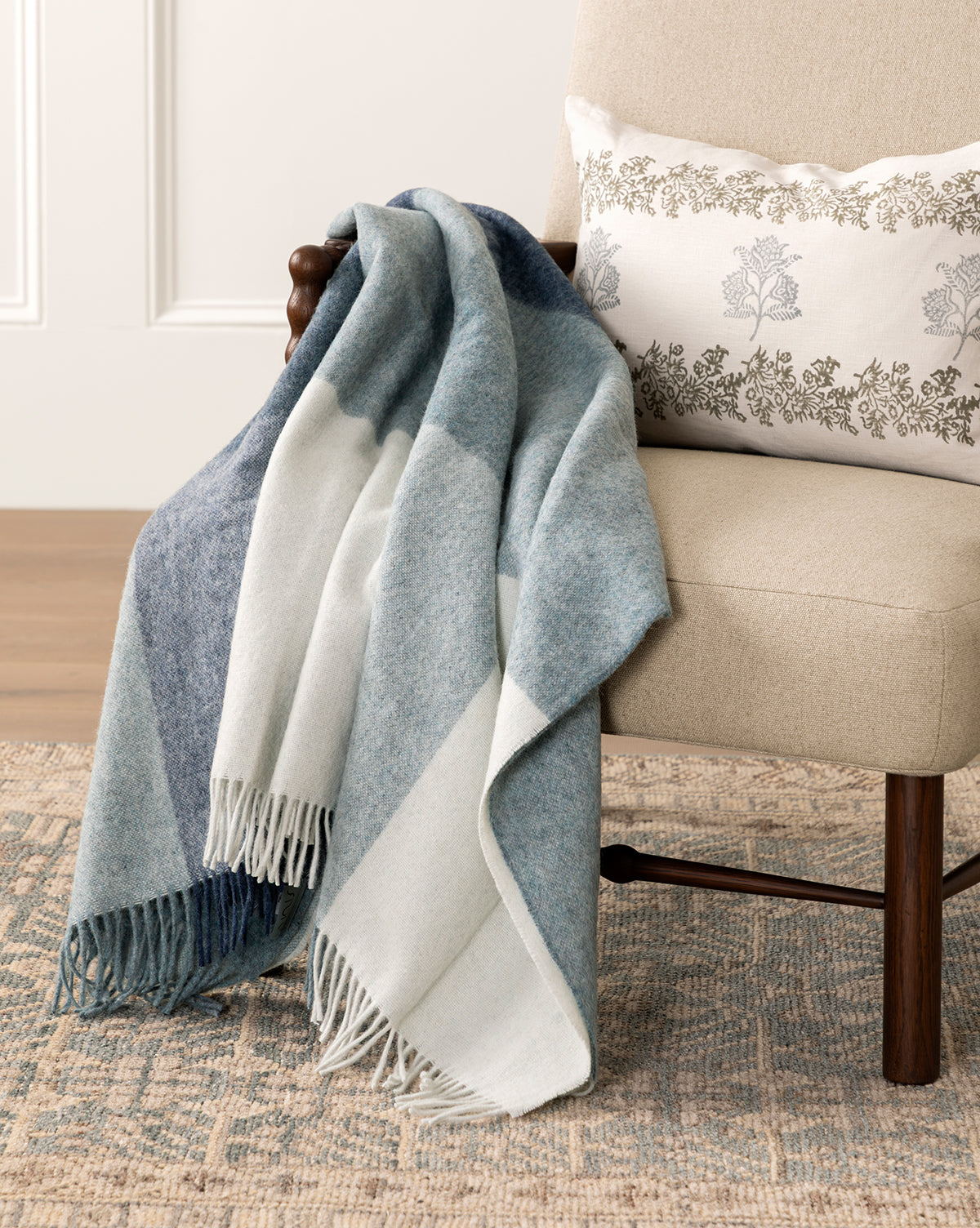 The Abner Throw, a soft blue plaid blanket made from recycled alpaca wool, drapes elegantly over a beige chair with a white floral pillow on a patterned rug—a cozy, eco-friendly accent for stylish comfort, by McGee & Co.