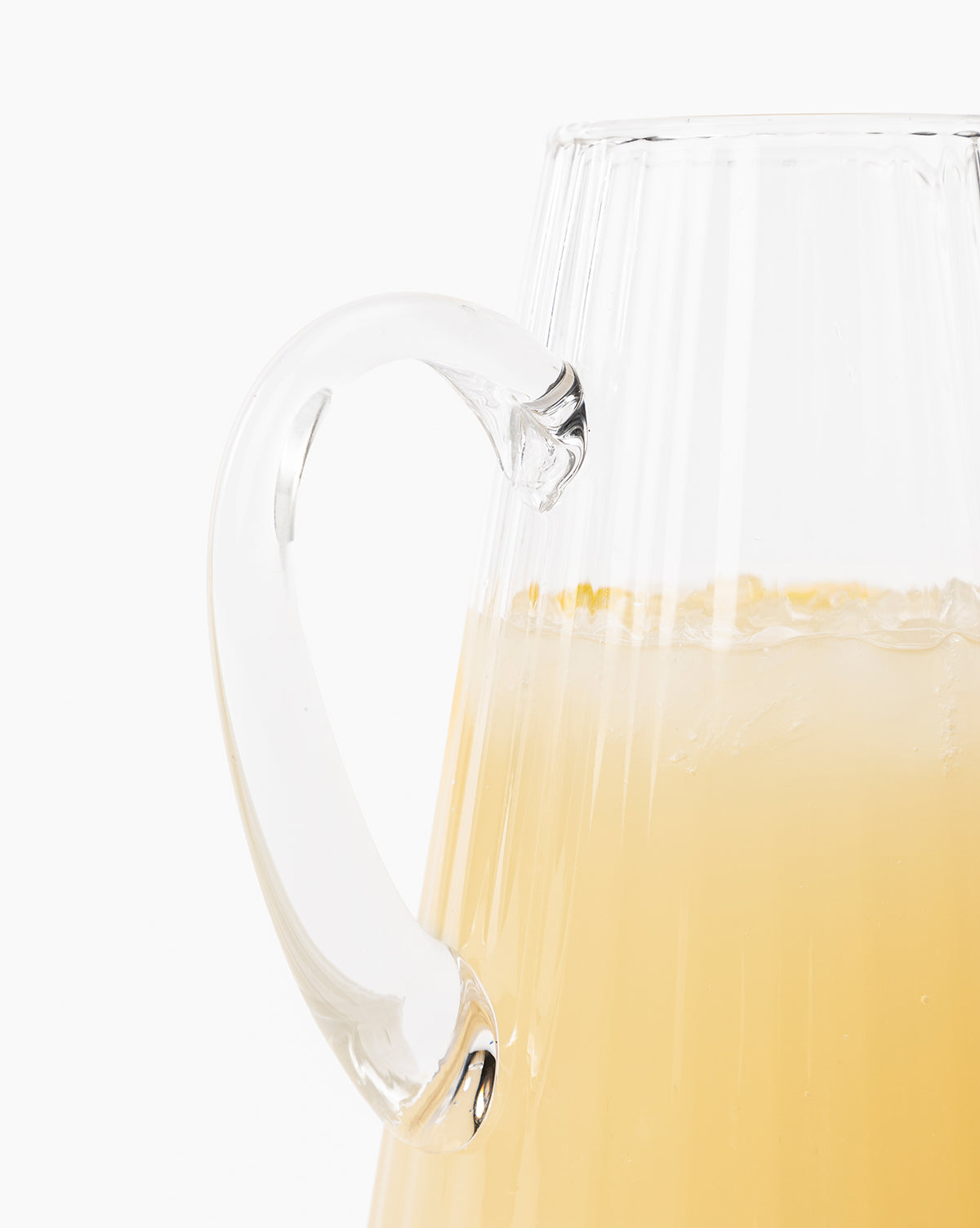 Close-up of the Adonis Ribbed Glass Pitcher filled with a pale yellow drink and ice, showcasing its textured surface and curved handle—ideal for stylish kitchen décor against a white backdrop, by McGee & Co.
