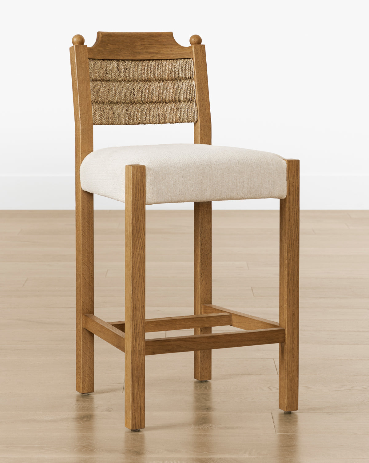 The Makerspalm Alberta Counter Stool has a woven seagrass rope backrest, beige cushioned seat, and honey oak legs, shown on a light wood floor against a white wall—McGee & Co.