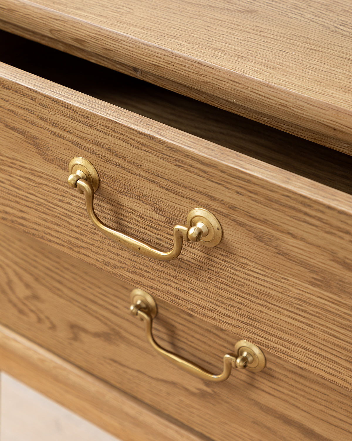 A close-up of two wooden drawers with brass handles on the Alessandra Nightstand, featuring a natural light oak finish, by McGee & Co. The top drawer is slightly open, revealing a dark gap, while the bottom drawer stays closed.