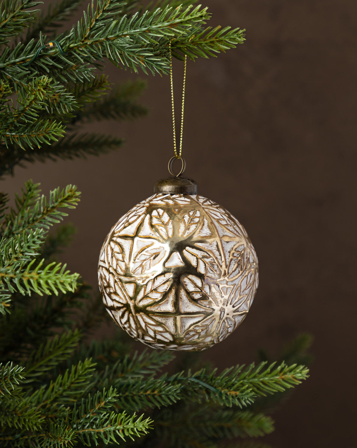 The Alessandro Glass Ornament, featuring an intricate gold pattern, hangs from a green pine branch against a neutral backdrop, bringing timeless charm to your holiday decor, by McGee & Co.