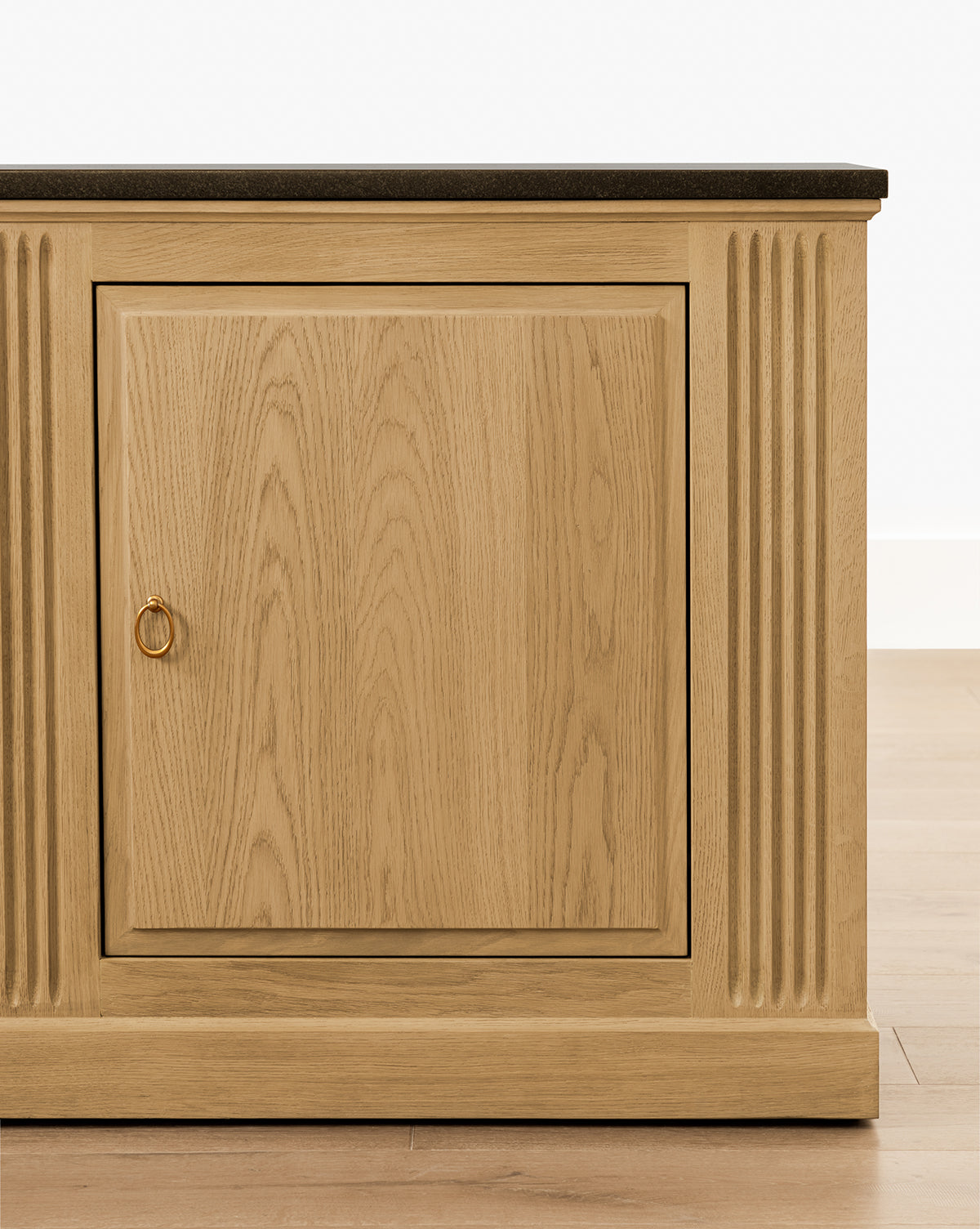 The Ambrose Sideboard features vertical grooves on the sides, a single central door with a round metal pull, and a dark countertop, by McGee & Co. It’s shown on a light wooden floor against a white wall.