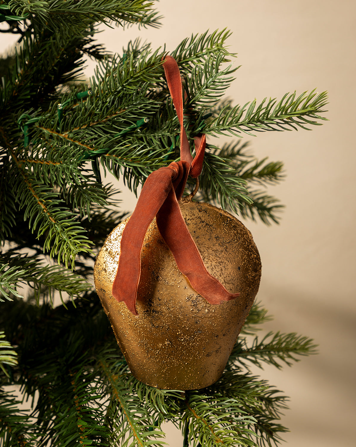 The Antique Gold Metal Bell with Velvet Ribbon hangs from a green Christmas tree branch against a neutral background, adding an elegant touch to your holiday décor, by McGee & Co.