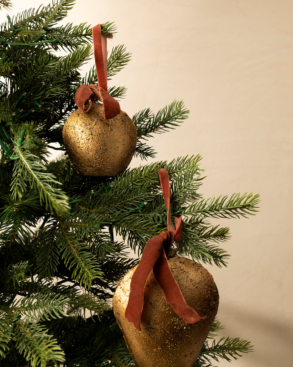 Two Antique Gold Metal Bell with Velvet Ribbon ornaments hang on a green Christmas tree, bringing classic holiday décor to a plain beige background, by McGee & Co.