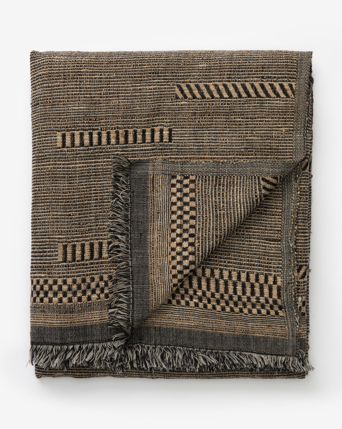 The Anwar Woven Throw is a cozy, elegant blanket with fringed edges, featuring earthy striped and checkerboard patterns. A flipped corner displays both sides of the design, showcasing its refined texture and style.