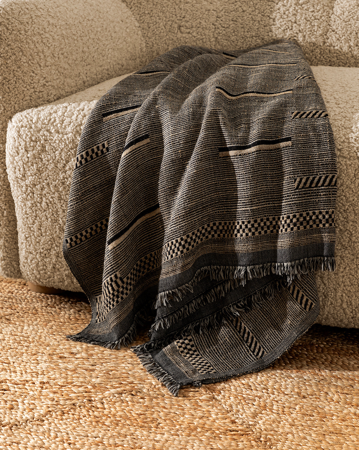 The Anwar Woven Throw, with its dark pattern and fringed edges, is draped over the arm of a textured beige sofa, complemented by a woven rug underneath.