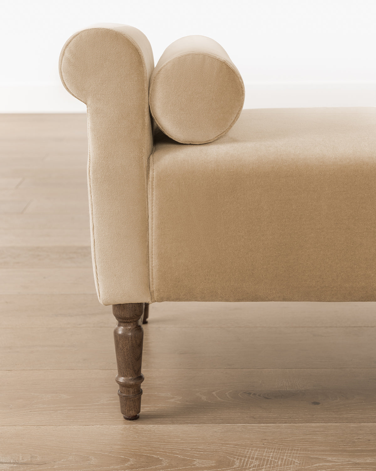 The Asher Bench, featuring beige striped linen upholstery and bolster pillows, rests on turned wooden legs atop a light wood floor with a white wall backdrop, by McGee & Co.