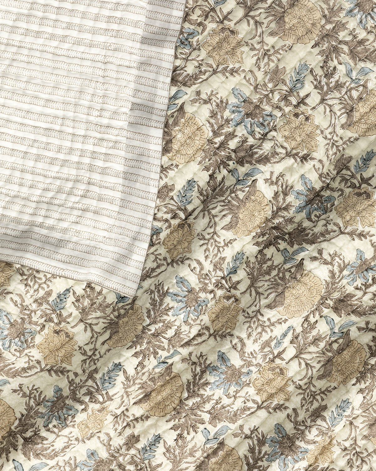 A cream-colored, ribbed blanket is partially draped over the Audrey Tan Block Print Quilt, which features beige, brown, and muted blue floral and leaf motifs on a light background, by McGee & Co.