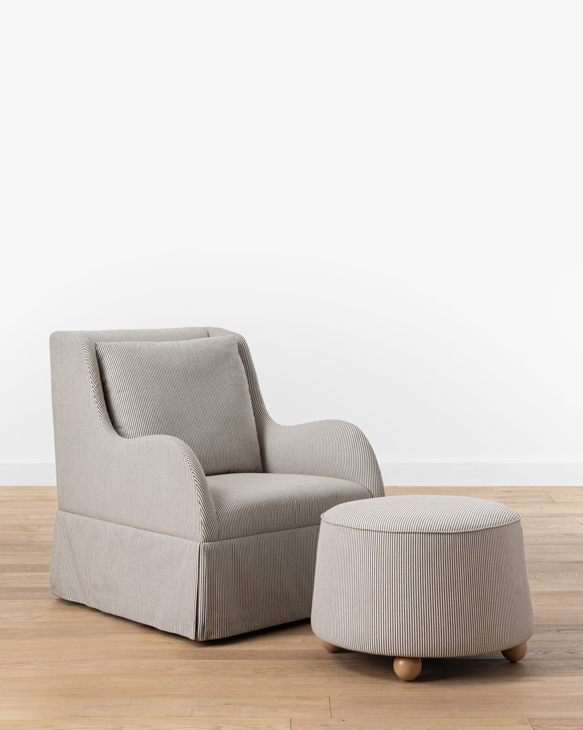 A plush, light-gray armchair with subtle vertical stripes is paired with the Barnaby Ottoman, both on ash wood legs, placed on a wooden floor against a plain white wall, by McGee & Co.