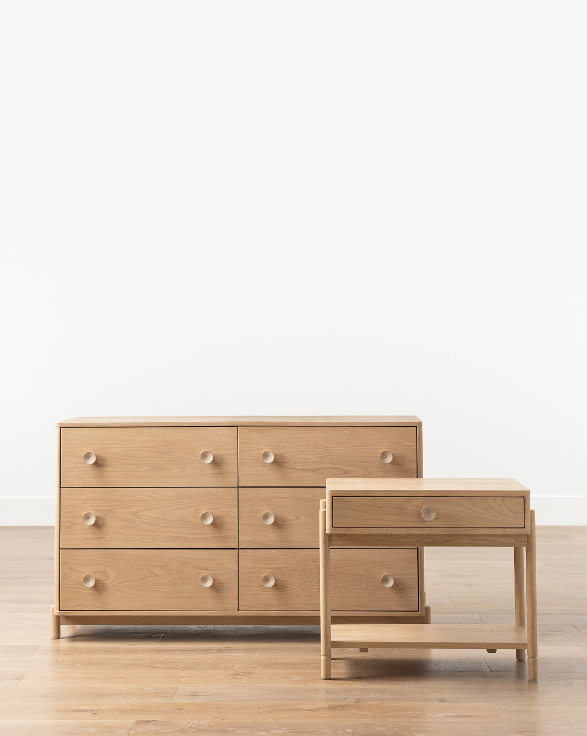 A six-drawer light wood dresser with round knobs stands next to the Baxter Nightstand, both featuring a transitional design and blending seamlessly on a wooden floor against a plain white wall, by McGee & Co.
