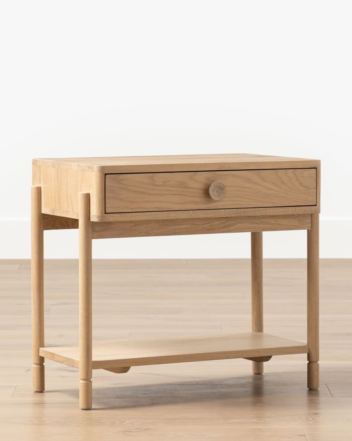 The Baxter Nightstand features a natural light oak finish, round legs, a single drawer with a round knob, and a lower open shelf. Its transitional design makes it an ideal bedroom piece, especially against white walls, by McGee & Co.