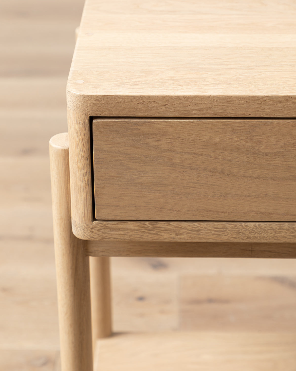 Close-up of the Baxter Nightstand shows its light oak finish, single drawer, and rounded legs on a matching floor. The minimalist design with smooth lines is perfect for transitional bedroom spaces, by McGee & Co.