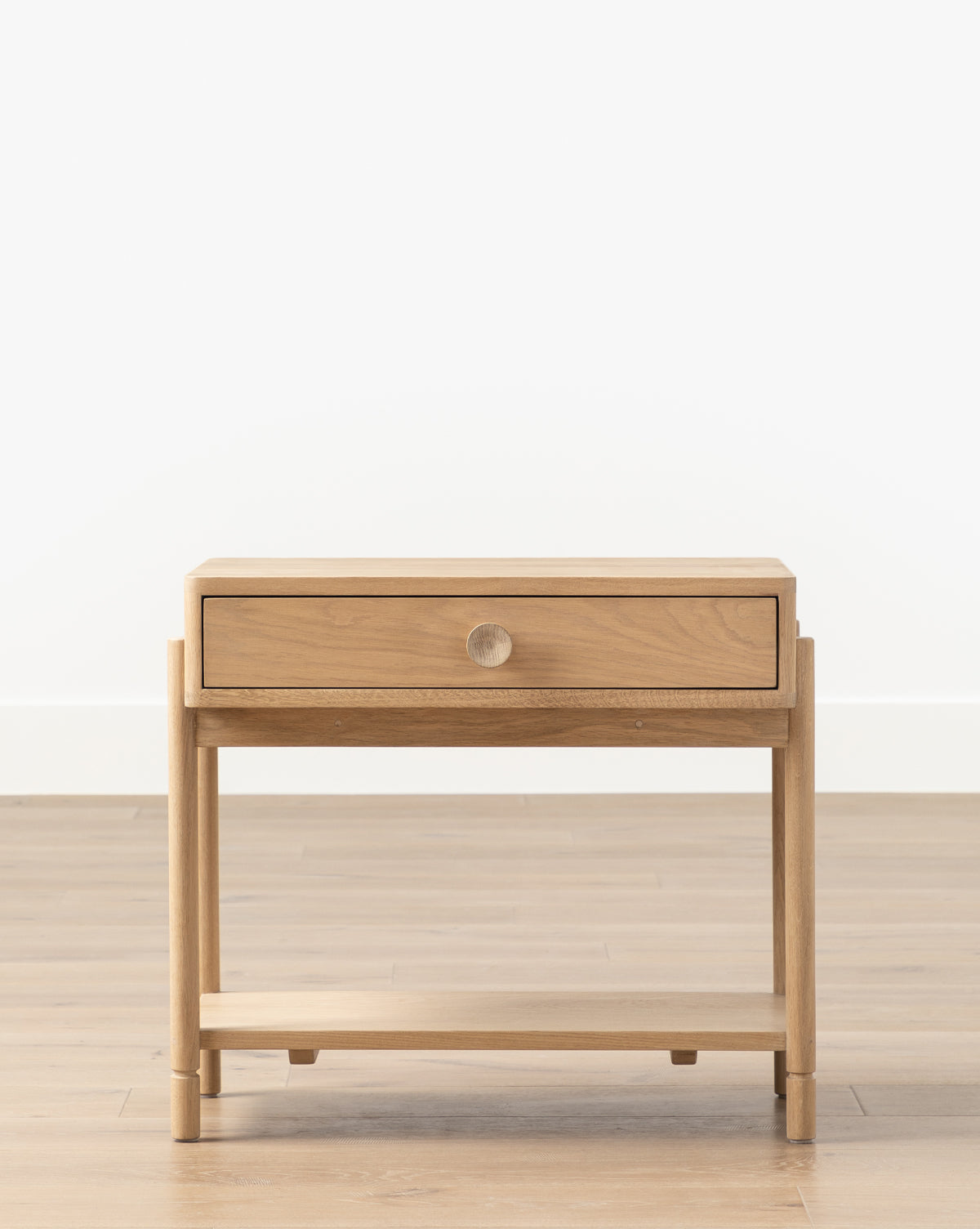 The Baxter Nightstand features transitional design in oak, a single drawer with a round knob, and an open lower shelf. Shown on a wood floor against a plain white background, it’s an elegant piece of bedroom furniture by McGee & Co.