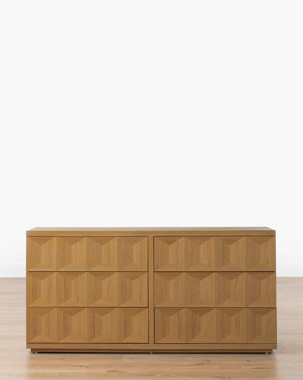 The Beacon 6-Drawer Dresser features a geometric 3D pattern on its fronts. Crafted from white oak, it stands elegantly on a wooden floor against a plain white wall, adding a modern touch to the bedroom, by McGee & Co.