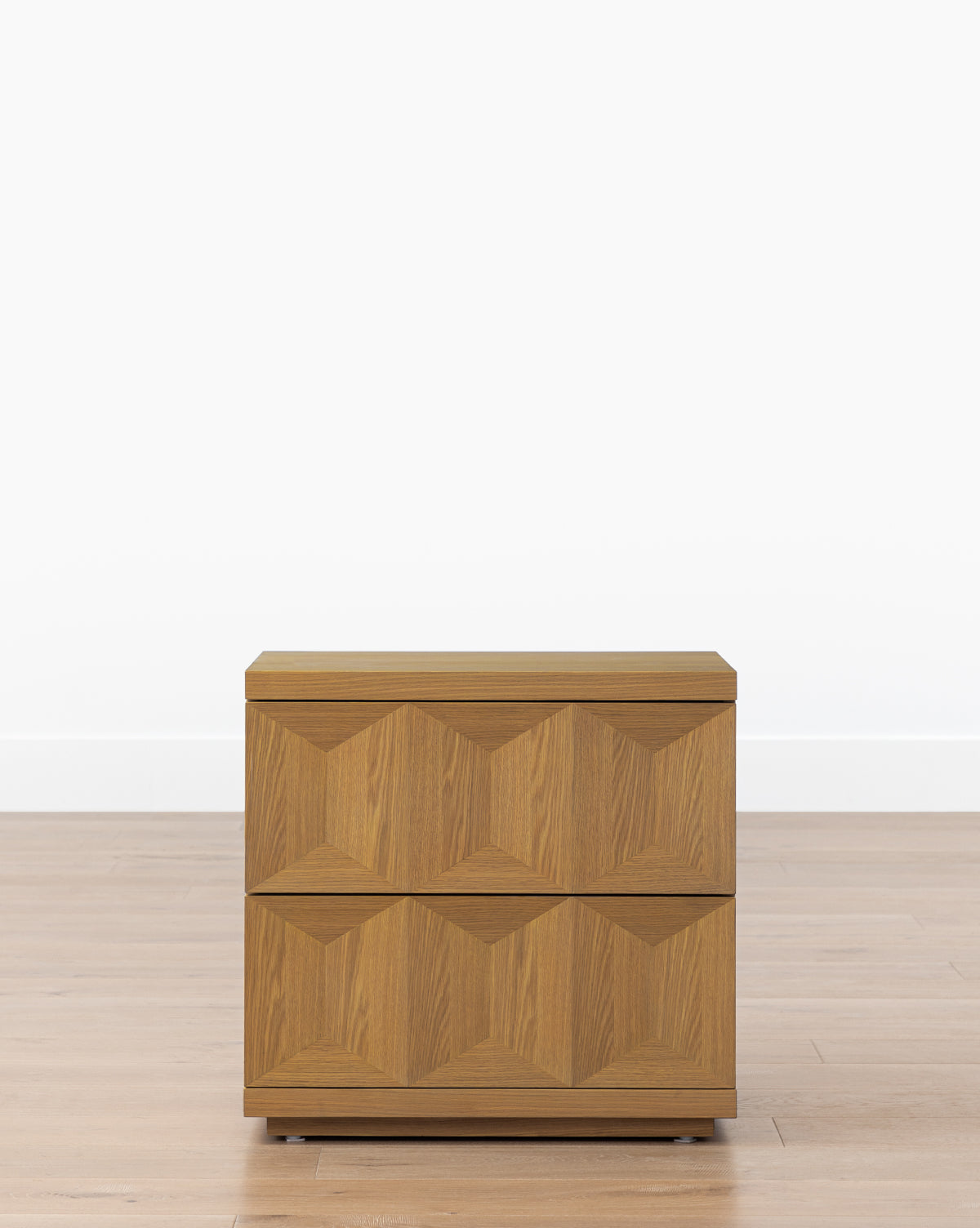 The Beacon Nightstand, made from white oak, features two drawers with a geometric textured front, offering stylish bedroom storage, by McGee & Co. It sits on a wooden floor against a plain white wall for a modern look.
