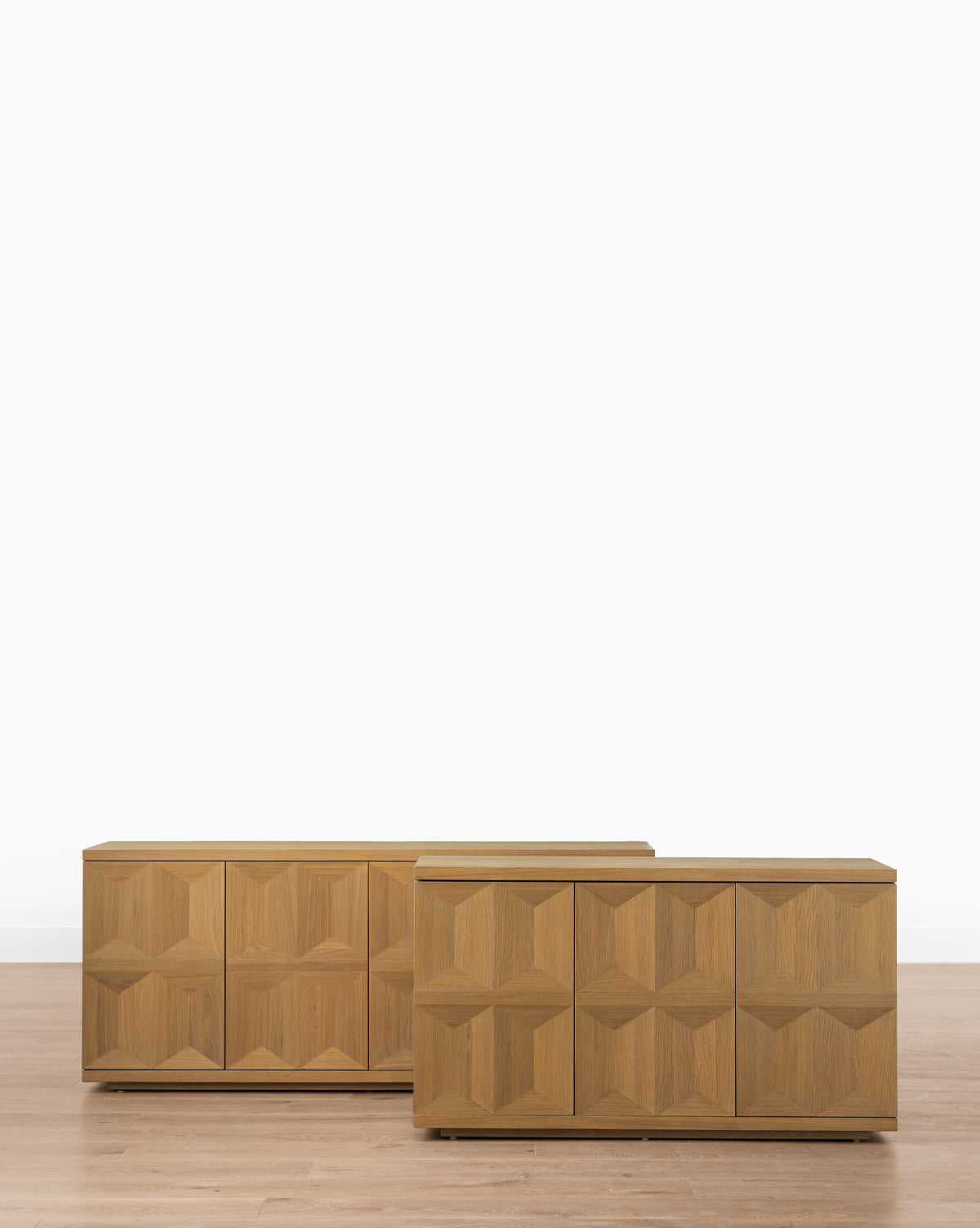 Two Beacon Sideboards with geometric, textured fronts and a neutral finish stand on a light wood floor by a white wall. One is slightly smaller and placed in front of the other, providing stylish and spacious storage by McGee & Co.