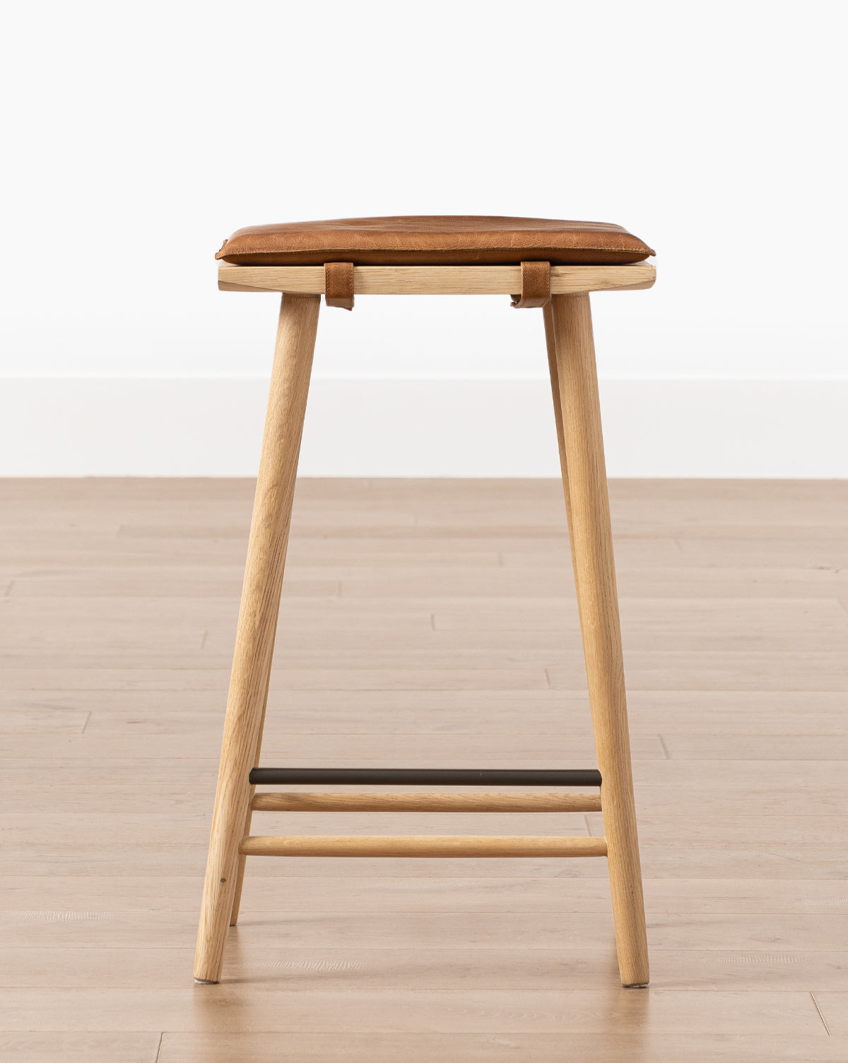 The Beau Counter Stool features a modern wooden frame, light brown leather seat, angled legs, and metal footrest bar, shown on a light wood floor with a white background by McGee & Co.